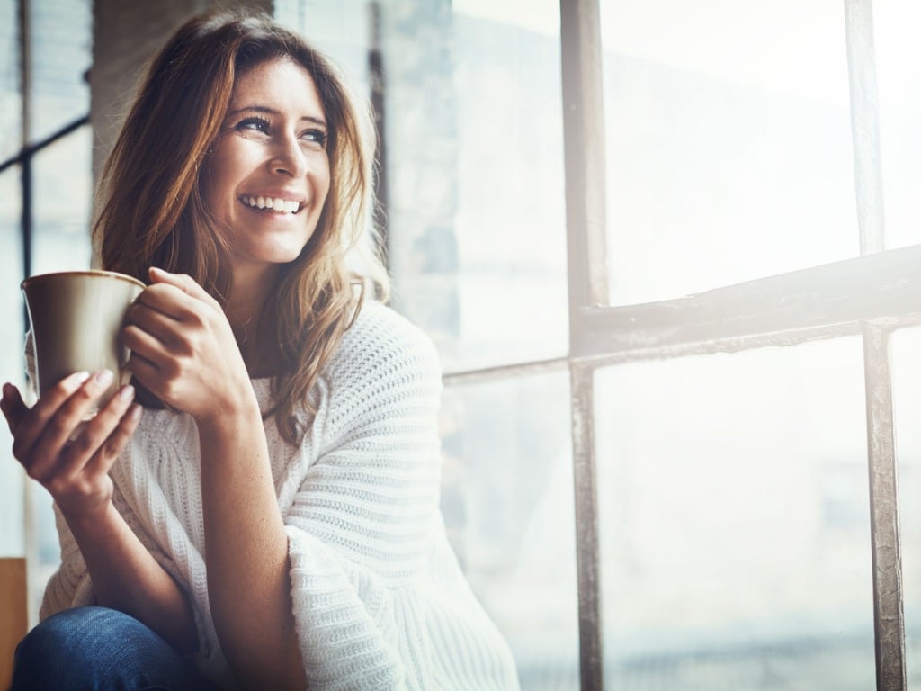 femme souriante et confiante, qui boit une boisson chaude et qui regarde par la fenêtre, son regard se projette vers l'avenir